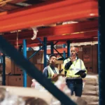 Warehouse staff in high-visibility vests inspecting inventory and reviewing stock information using a tablet in a storage facility.