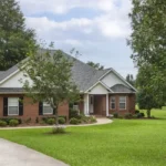Traditional style brick house with black shutters, situated on a large green yard with mature trees.